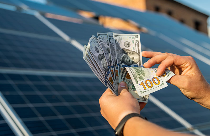 Person holding stacks of $100 bills in front of a solar panel